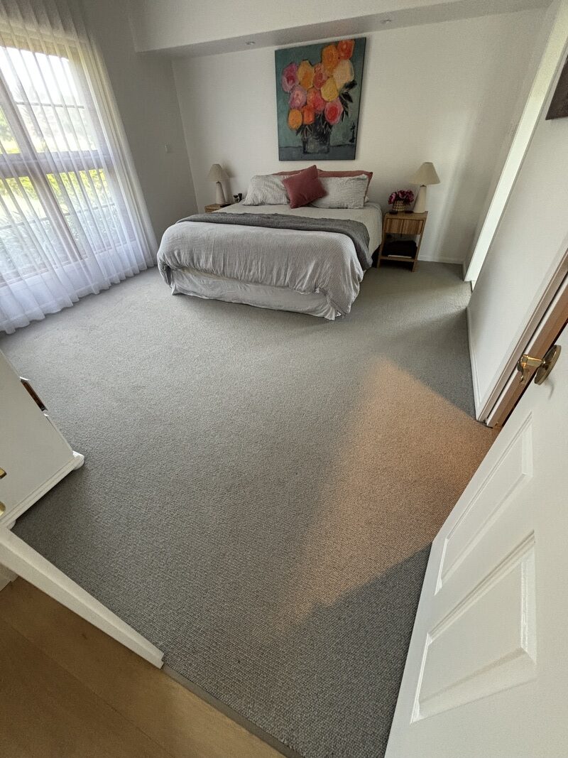 Bedroom with freshly cleaned carpet, bright natural light from window, decorative flower painting on wall, and neatly arranged bedding.