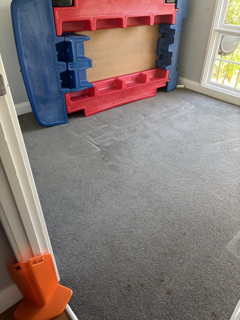 Bedroom carpet with visible stains and soiling from a child's accident, featuring a red and blue play structure in the background and an orange doorstop in the foreground.